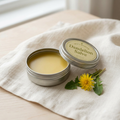 Open silver tin of Dandelion Salve on ivory linen beside a dandelion flower
